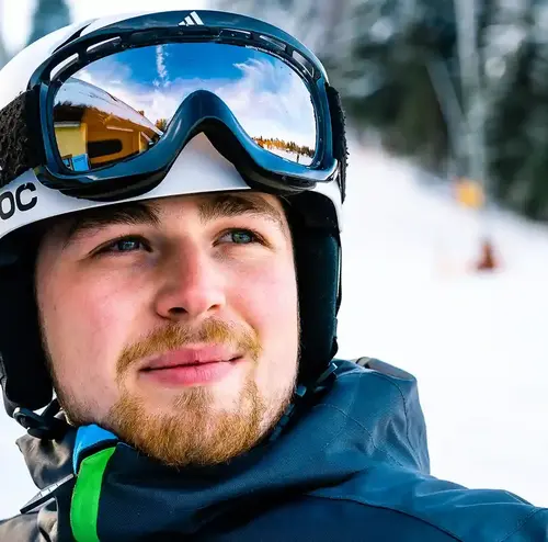 Ein junger Mann steht auf der verschneiten Piste und schaut in die Ferne. er trägt Skiausrüstung sowie einen Helm und eine Skibrille.