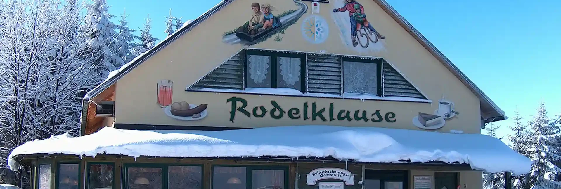 Auf dem Bild sieht man ein verschneites Haus, die Rodelklause. Dort gibt es leckere Snacks und warme Getränke perfekt nach verschneiten Abfahrten. Auch im Sommer gibt es dort leckere Snacks sowie erfrischende Getränke. Besuchen sie uns gerne am Erlebnisberg in Altenberg.