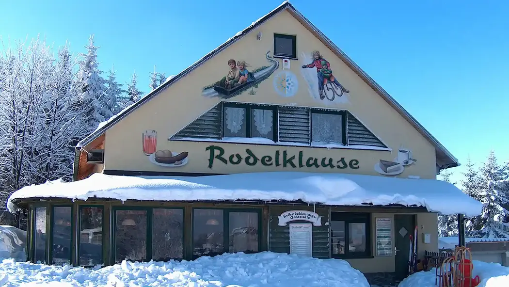 Auf dem Bild sieht man ein verschneites Haus, die Rodelklause. Dort gibt es leckere Snacks und warme Getränke perfekt nach verschneiten Abfahrten. Auch im Sommer gibt es dort leckere Snacks sowie erfrischende Getränke. Besuchen sie uns gerne am Erlebnisberg in Altenberg.