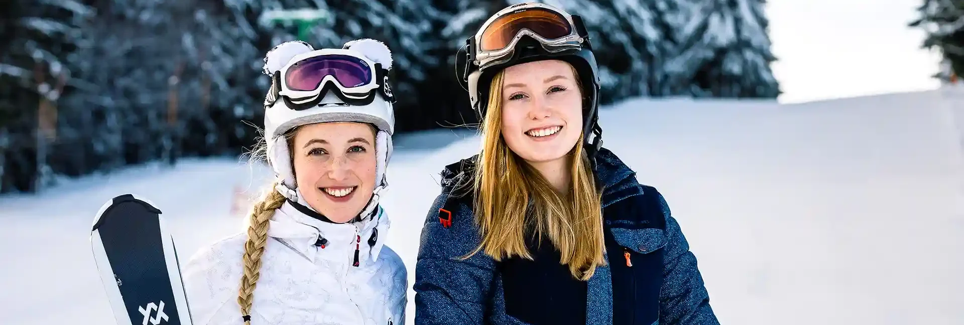 Zwei junge Damen stehen in Skiklamotten lächelnd auf einer verschneiten Piste. Im Hintergrund ist ein kleines Waldstück.