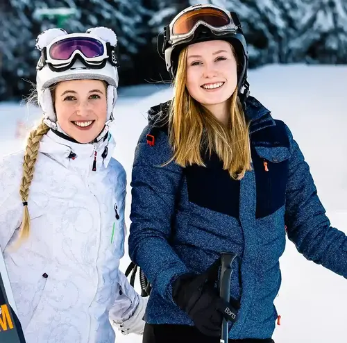 Zwei junge Damen stehen in Skiklamotten lächelnd auf einer verschneiten Piste. Im Hintergrund ist ein kleines Waldstück.