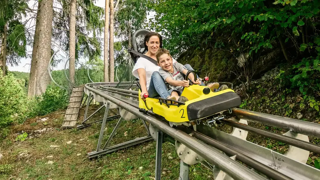 Eine Frau und ihr Sohn fahren zusammen mit der Sommerrodelbahn. Sie fahren durch ein Waldstück und man sieht die grünen Bäume. Beide haben sichtlich Spaß, die Mutter hält den Bremshebel, während der Sohn sich am Schlitten festhält. 