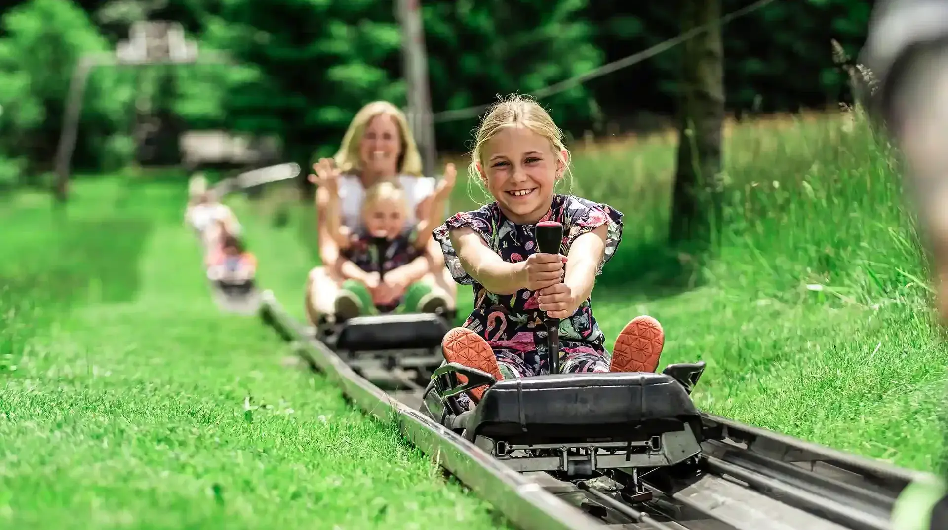 schulklassen-im-sommer-altenberg-erzgebirge-sachsen-09.webp Ein kleines Mädchen fährt lachend mit der Sommerrodelbahn vor einer Kulisse mit grünen Bäumen im Erzgebirge