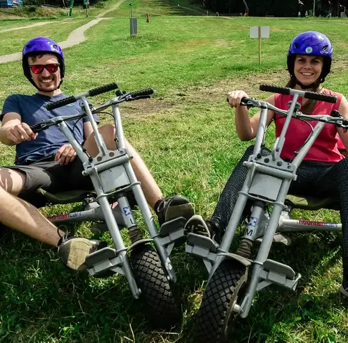 vereine-altenberg-erzgebirge-sachsen-05.webp Zwei Erwachsene stehen mit ihren Mountaincarts auf einer grünen Wiese am Erlebnisberg Altenberg.