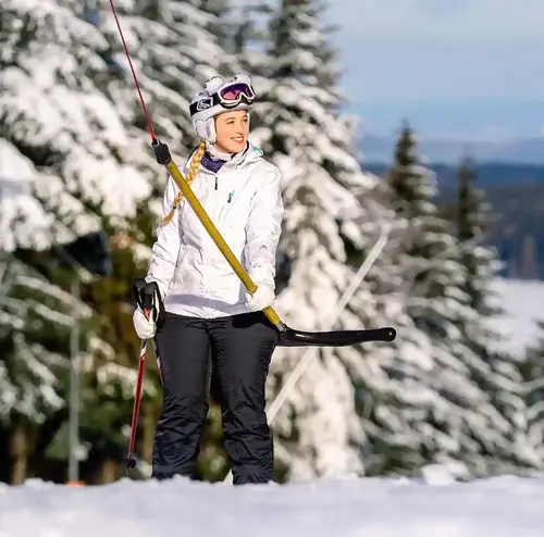 Eine junge Frau fährt auf Skiern und in Skiausrüstung mit dem Schlepplift den Berg hinauf. Im Hintergrund sieht man ein verschneites Waldstück.
