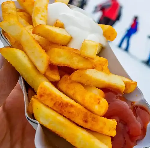 Auf der verschneiten Piste hält eine Person eine Portion Pommes mit Ketchup und Mayonnaise. Im Hintergrund erkennt man Ski fahrende Personen. Entdecken sie die Rodelklause mit Snacks und Getränken am Erlebnisberg in Altenberg.