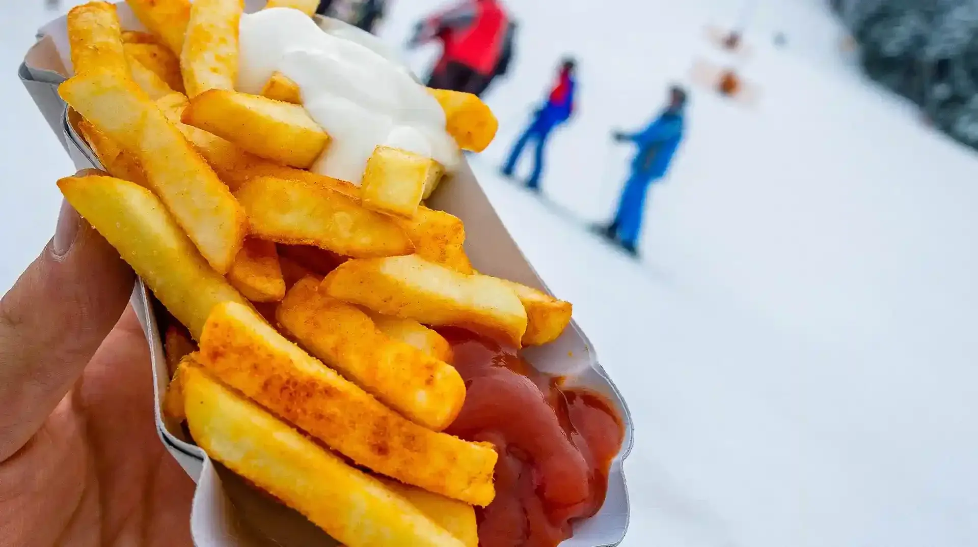 Auf der verschneiten Piste hält eine Person eine Portion Pommes mit Ketchup und Mayonnaise. Im Hintergrund erkennt man Ski fahrende Personen. Entdecken sie die Rodelklause mit Snacks und Getränken am Erlebnisberg in Altenberg.