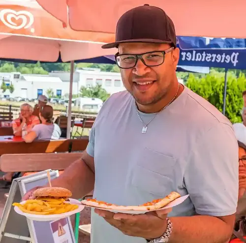 Ein junger Mann steht im Biergarten der Rodelklause und hat einen Burger und eine Pizza in der Hand. Viele Leute im Hintergrund genießen das schöne Wetter. Entdecken sie leckere Snacks und Getränke in der Rodelklause am Erlebnisberg in Altenberg.