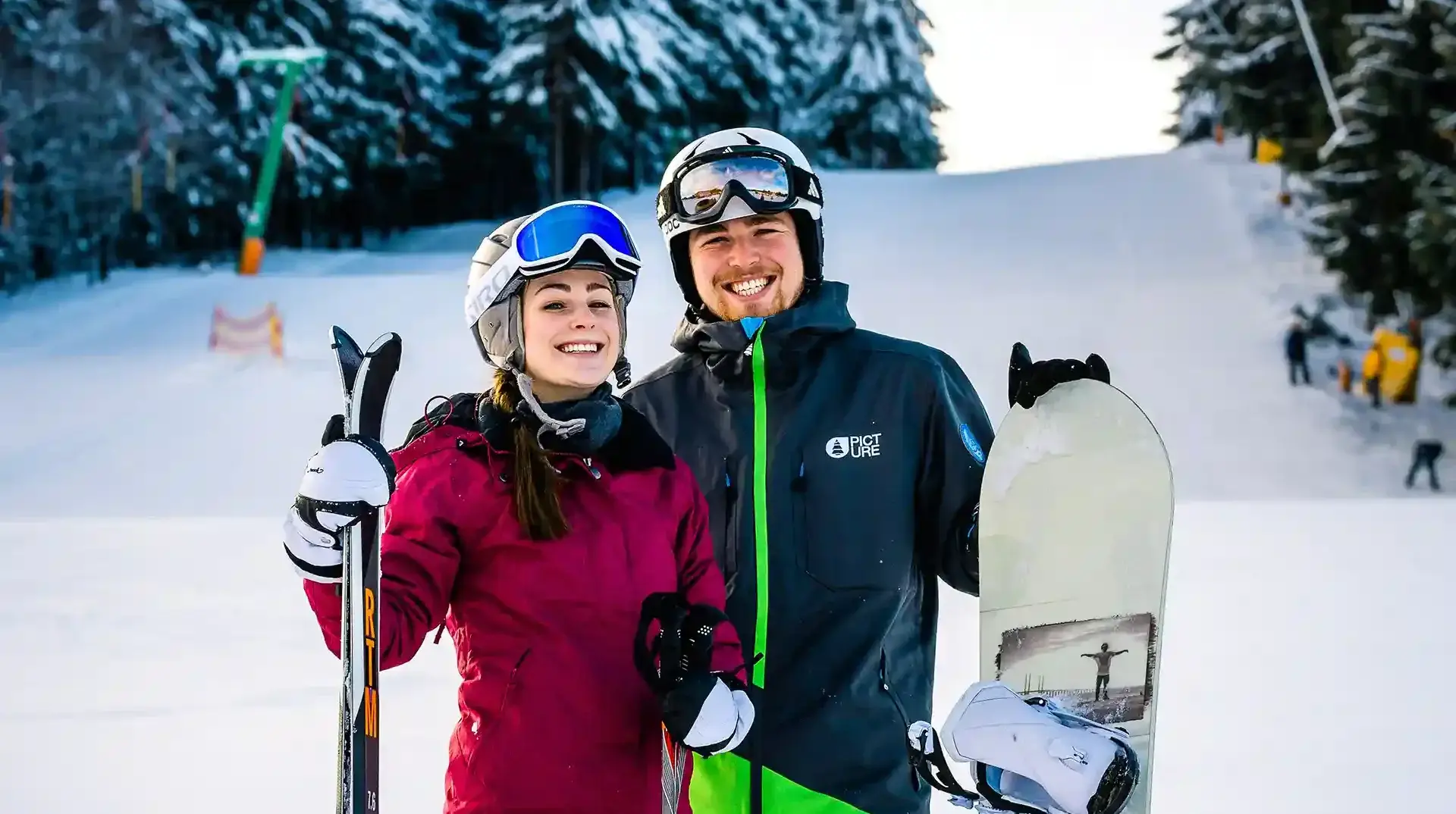 Ein junges Paar steht lächelnd auf der verschneiten Piste. Sie tragen ihre Skiausrüstung und im Hintergrund erkennt man einen Lift und ein kleines Waldstück.