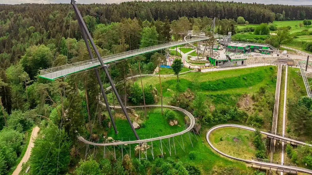 Man sieht eine frei schwebende Brücke, den Skywalk. Unter dem Skywalk verläuft eine Strecke für den Alpine Coaster. Alles ist umringt von Bäumen und im Hintergrund sieht man noch Gebäude stehen. 
