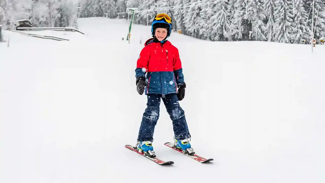 Ein grinsender Junge steht mit Skiern auf der verschneiten Piste. In Hintergrund sieht man noch den Skilift.