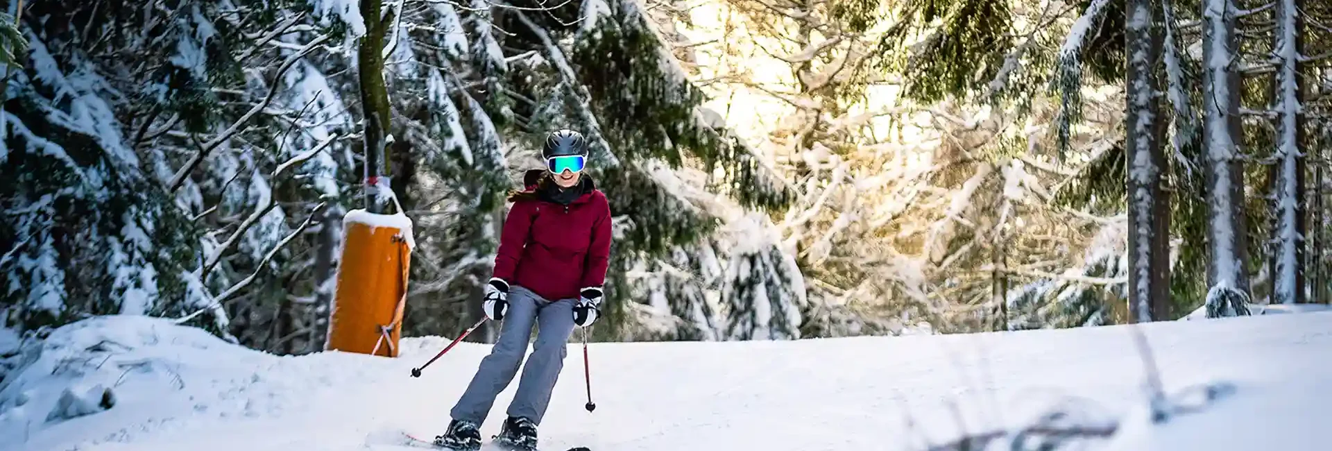 Eine Frau mit Winterklamotten und Skistöcken, fährt entspannt die Piste entlang. Sie fährt durch einen verschneiten Wald und im Hintergrund sieht man einzelne Sonnenstrahlen.