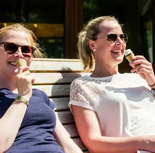busreisen-altenberg-erzgebirge-sachsen-12.webp Zwei Frauen essen fröhlich ein Eis auf einer Bank am Erlebnisberg Altenberg.