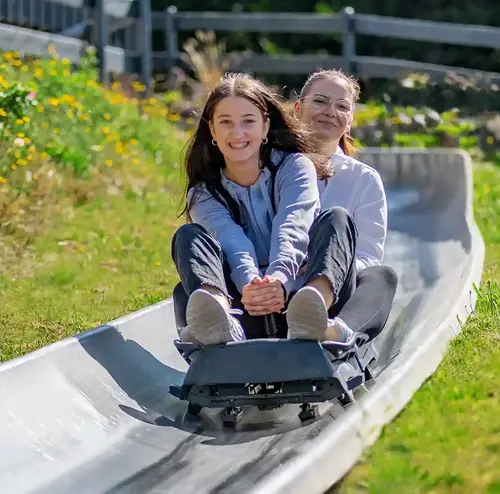 schulklassen-im-sommer-altenberg-erzgebirge-sachsen-16.webp Zwei junge Frauen fahren grinsend mit der Sommerrodelbahn am Erlebnisberg Altenberg.