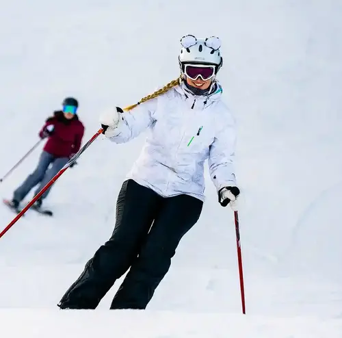 Zwei junge Damen sind auf einer Piste unterwegs. Die Piste ist verschneit und sie haben sichtlich Spaß auf den Skiern.