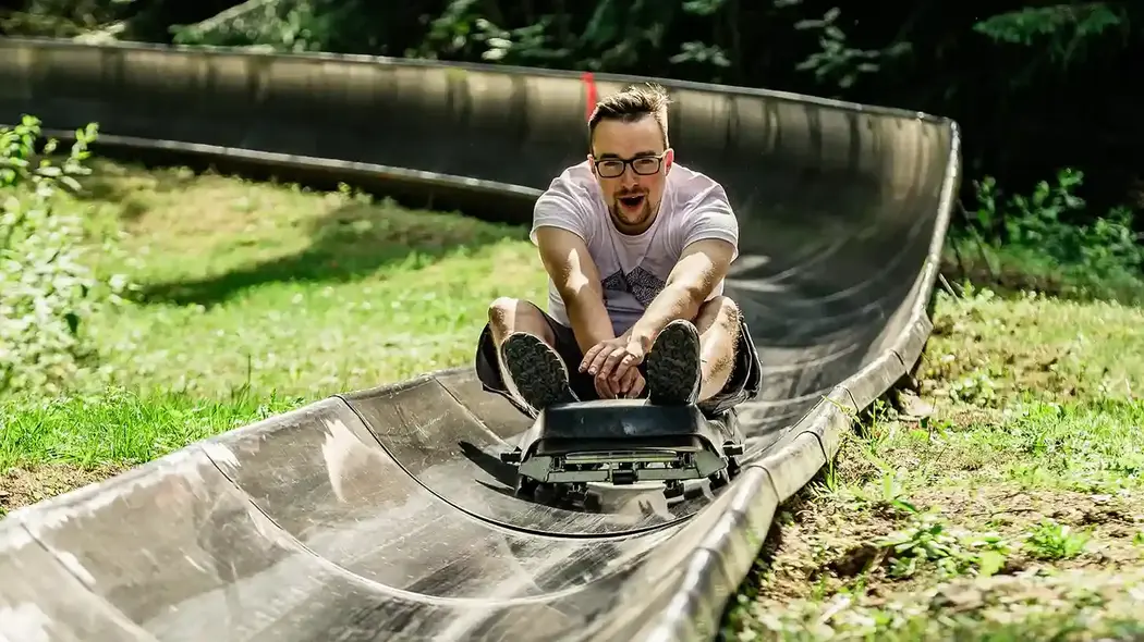 Ein Mann fährt im Sommer auf einer Sommerrodelbahn. Er fährt inmitten der Natur und hat sichtlich Spaß. Er hält sich am Schlitten fest.