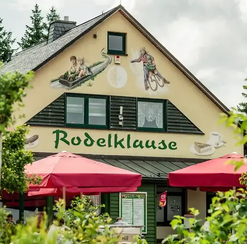 Man sieht ein Gasthaus im Hintergrund. Vor dem Haus und auch rundherum sind viele Bäume. Der Biergarten vor dem Gasthaus sieht sehr einladend aus. Entdecken sie leckere Snack, Speisen und Getränke in der Rodelklause am Erlebnisberg in Altenberg.