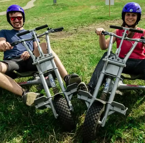 busreisen-altenberg-erzgebirge-sachsen-05.webp Ein junger Mann und eine junge Frau stehen lachend mit ihren Mountaincarts am Erlebnisberg Altenberg.