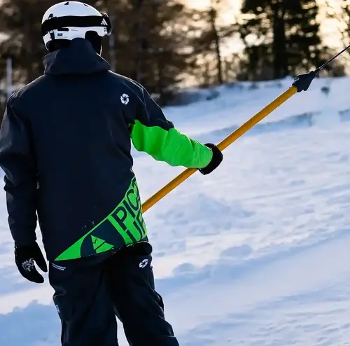 Man sieht eine Person von hinten, sie nutzt gerade den Skilift. Die Piste ist verschneit und im Hintergrund sieht man ein kleines Waldstück.