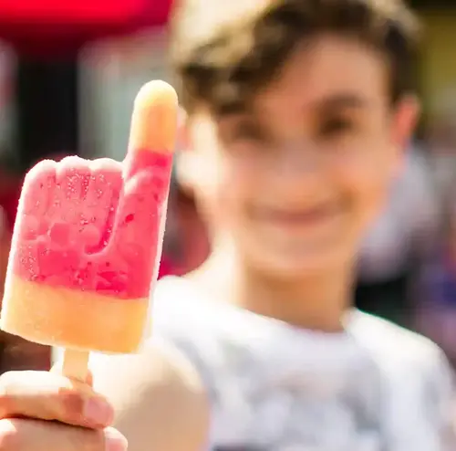Auf dem Bild ist ein Junge zu sehen, der ein handförmiges Eis isst. Man sieht ihn im Hintergrund verschwommen grinsen. Entdecken sie erfrischen Angebote in der Rodelklause am Erlebnisberg in Altenberg.