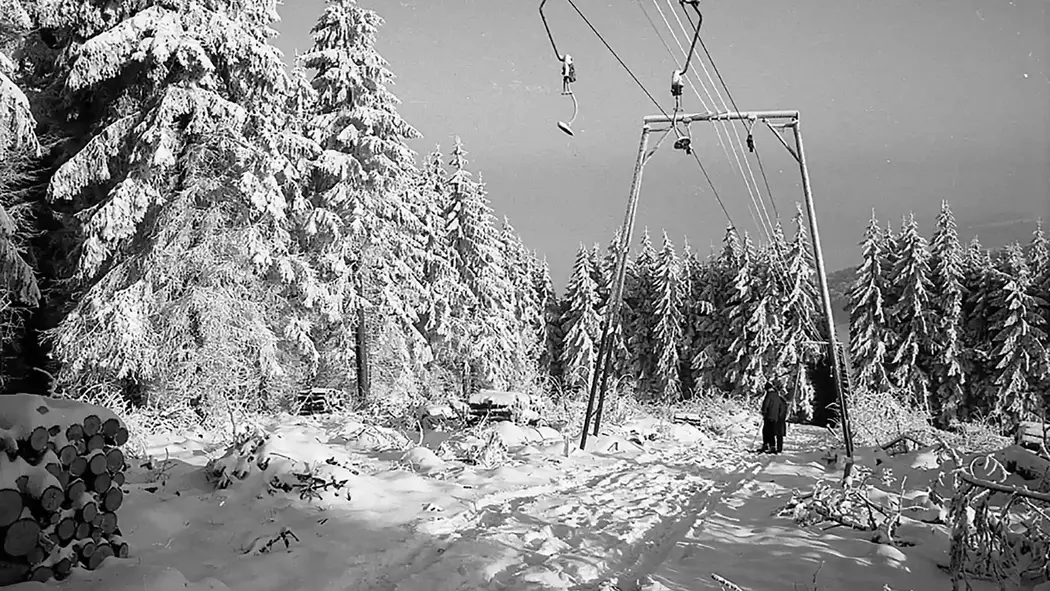Man sieht eine schwarz-weiß Aufnahme eines alten Skilifts von Josef Wiegand. Es liegt Schnee und um den Lift herum sind hohe Tannen. Unter dem Lift stehen zwei Personen auf Skiern.