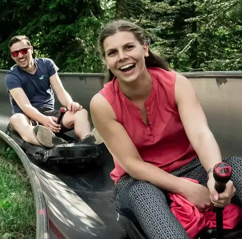 Ein junger Mann und eine junge Frau fahren mit einem lächeln die Sommerrodelbahn hinunter. Inmitten der schönen Natur. Entdecken sie spannende Attraktionen am Erlebnisberg in Altenberg.
