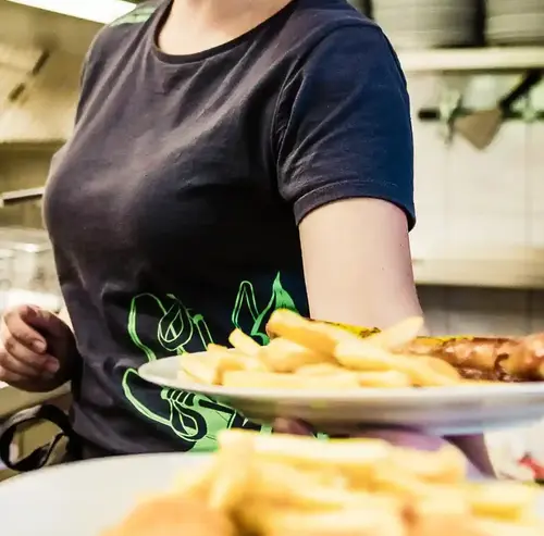 Eine junge Dame, die in der Küche arbeitet, hält 2 Teller in der Hand, mit Pommes, Wurst und Chicken Nuggets. Die Küche ist sehr sauber und die Speisen sehen ebenfalls gut aus. Entdecken sie leckere Snacks, Speisen und Getränke in der Rodelklause am Erlebnisberg in Altenberg.