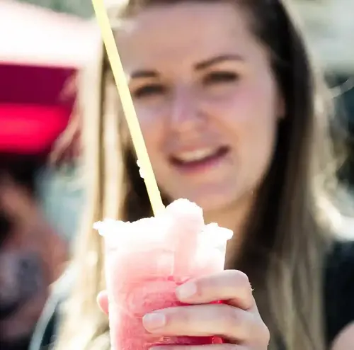 Eine junge Dame schlürft ein Slush-Eis und man sieht sie im Hintergrund verschwommen grinsen. Erleben sie erfrischende Momente und abkühlende Snacks und Getränke in der Rodelklause am Erlebnisberg in Altenberg.