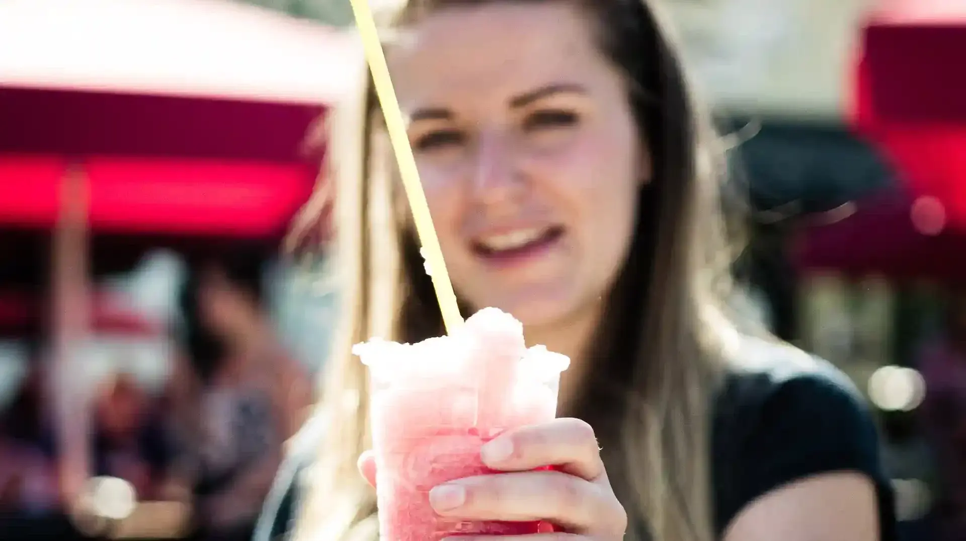 Eine junge Dame schlürft ein Slush-Eis und man sieht sie im Hintergrund verschwommen grinsen. Erleben sie erfrischende Momente und abkühlende Snacks und Getränke in der Rodelklause am Erlebnisberg in Altenberg.